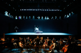 La gran orquesta del Teatro Real, el escenario desnuda con los personajes clave rodeando a La Regenta, y arriba el gran coro representando a la alta sociedad de la «beata Vetusta».